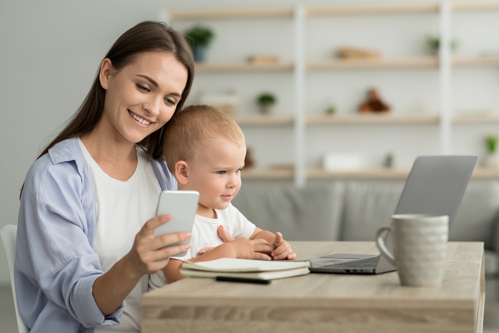 Remote Career. Young Mom With Baby On Hands Using Smartphone And Laptop